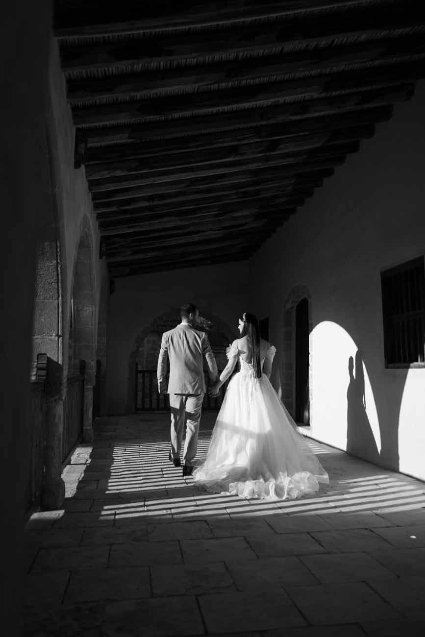 Bride and groom walking hand in hand through a sunlit corridor, in black and white