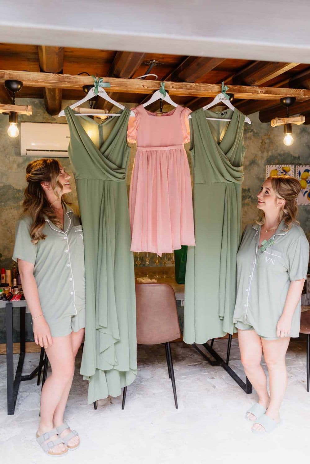 Bridesmaids in matching gray robes examine sage green and blush pink dresses hanging on wo