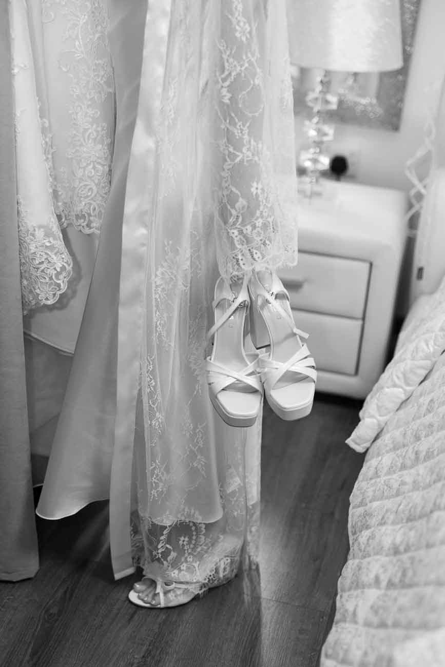 Black and white photo of elegant bridal heels hanging beside lace wedding gowns in a brida