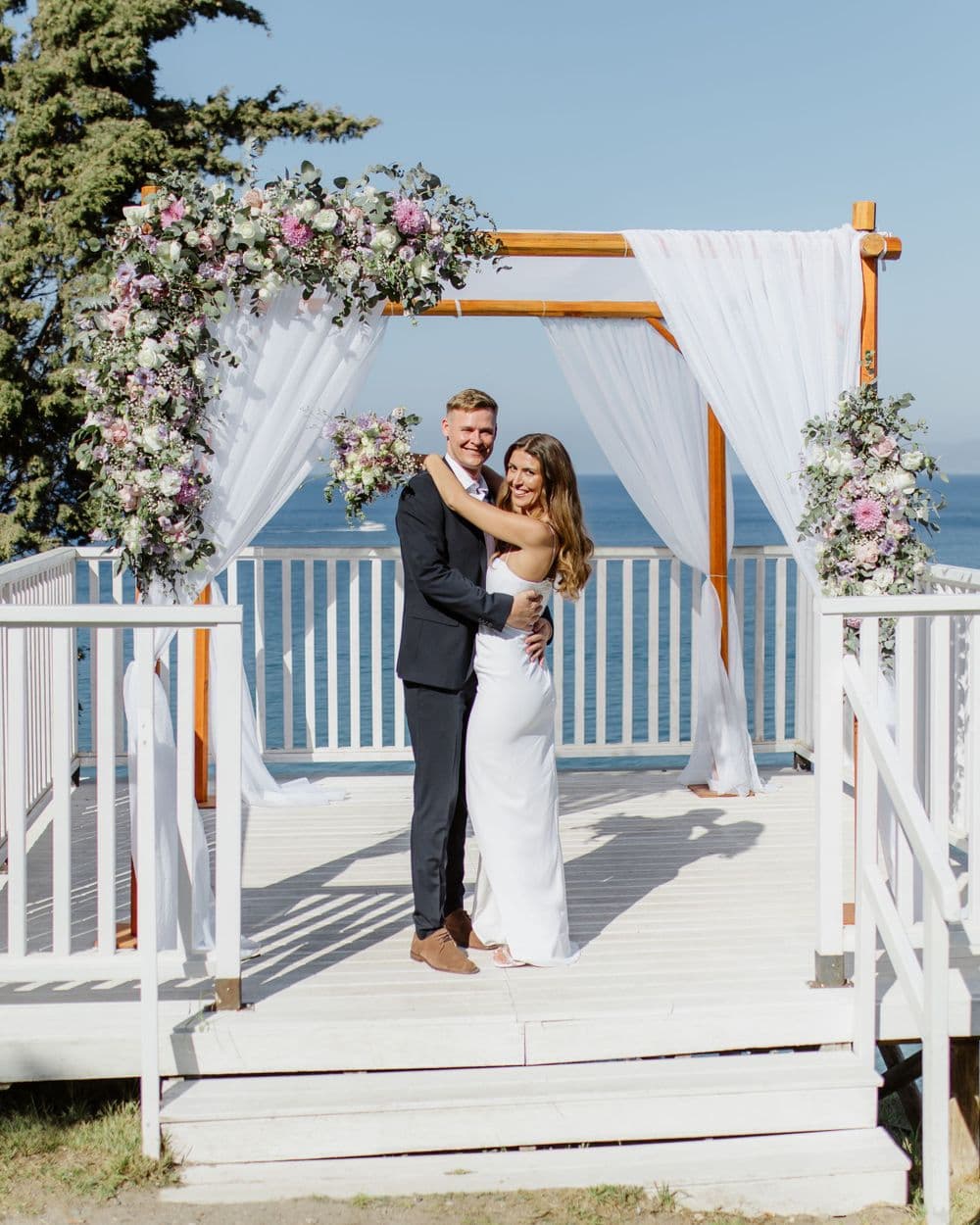 A romantic wedding ceremony takes place under a white draped arch decorated with pink and 