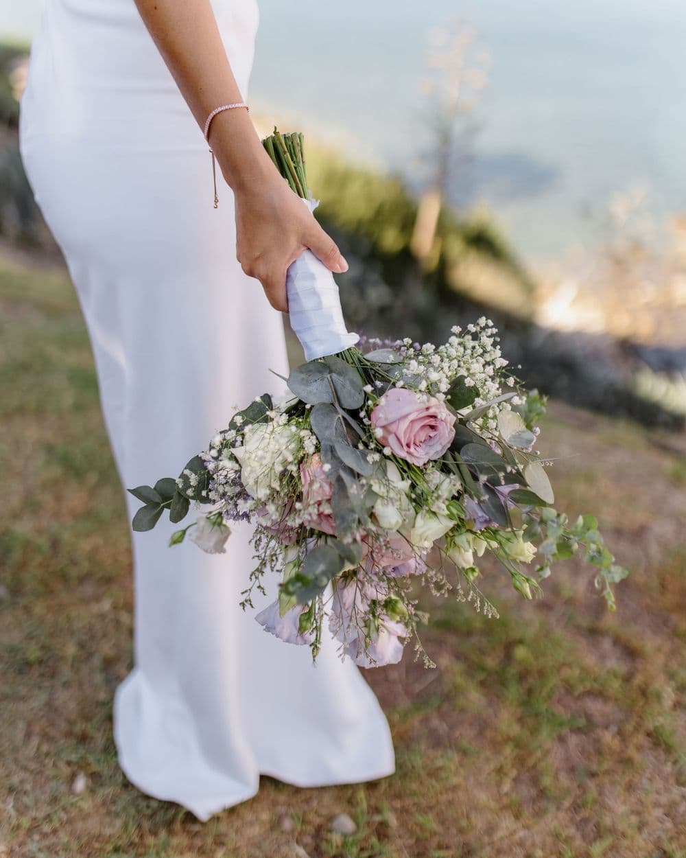 A delicate wedding bouquet with pink roses and baby’s breath held against a white dress ba