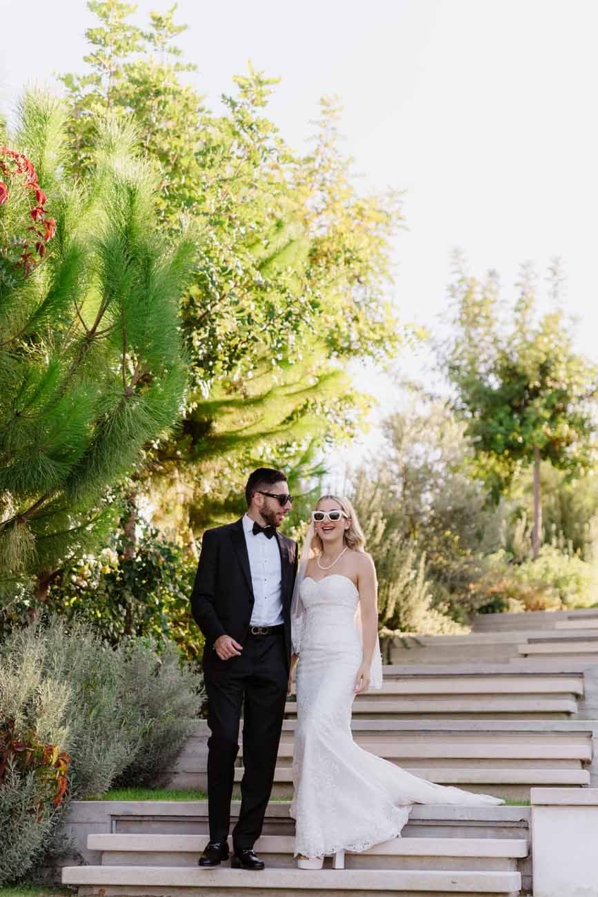 Bride and groom pose on outdoor steps surrounded by lush greenery wearing sunglasses on we