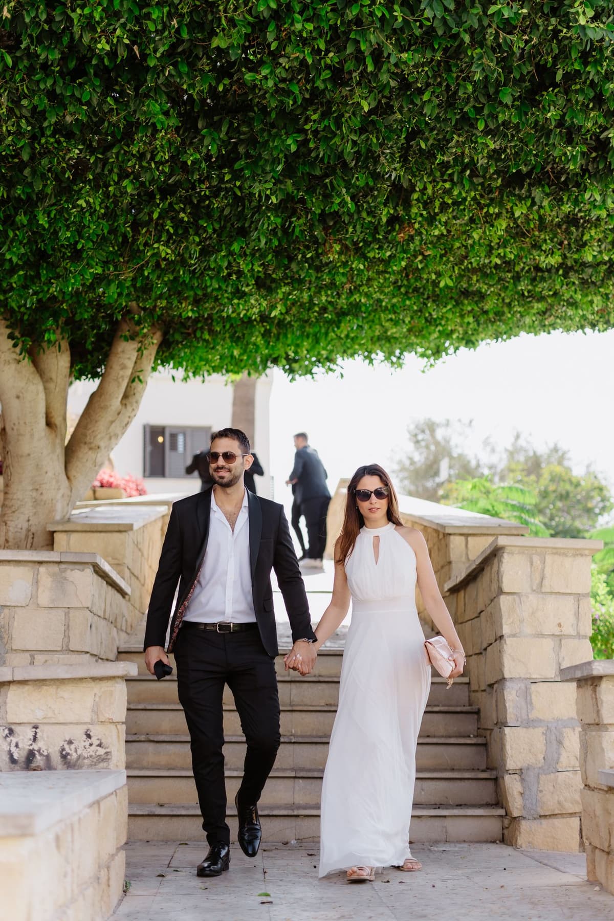 A couple descending stone steps together under lush green trees at an outdoor venue.