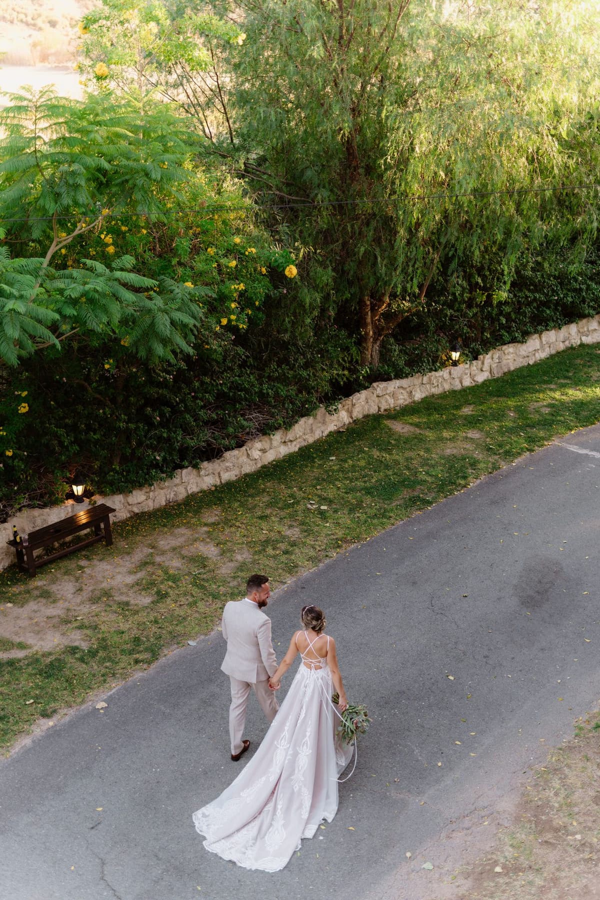 Bride and groom walk hand-in-hand down a quiet road, surrounded by greenery, captured from