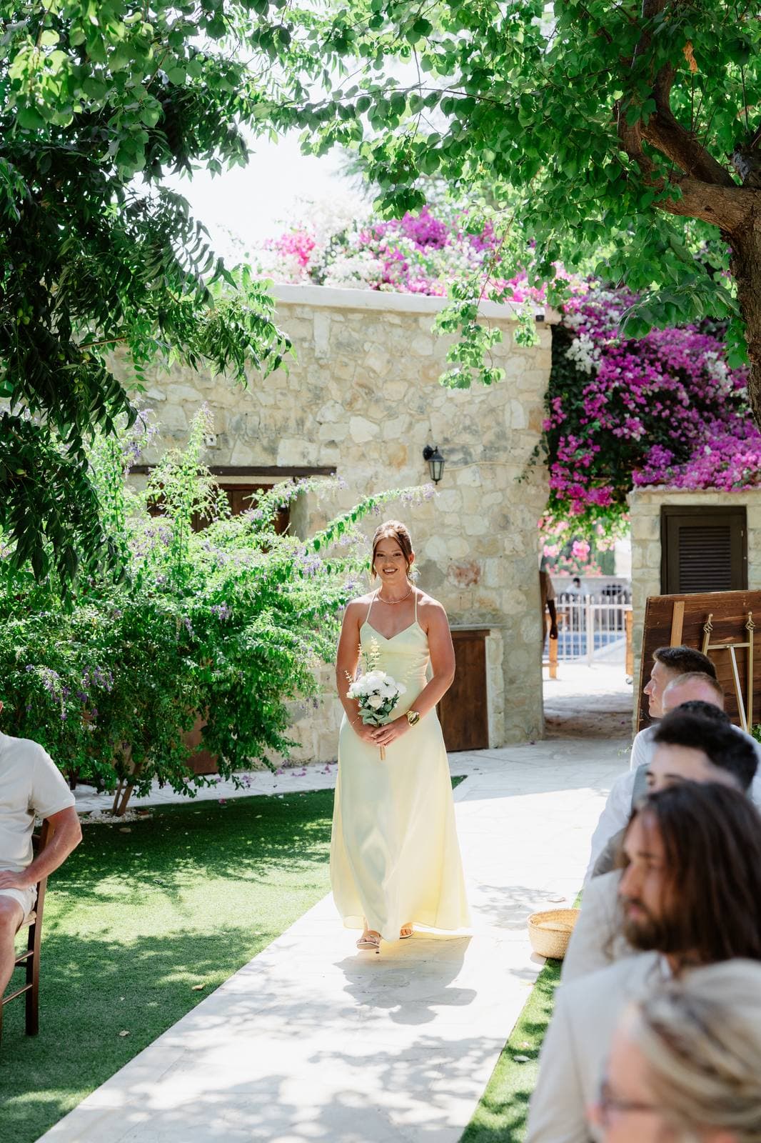 Hannah & Danial, a bridesmaid walking down the aisle under the bougainvillea